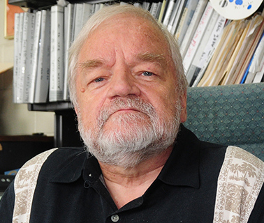 David, an older gentleman with silver hair and beard, sits in a chair and looks into the camera