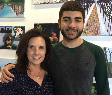 John, a young man with dark hair and beard poses with his Mom, Eileen. John has his arm around his mom’s shoulder and both smile